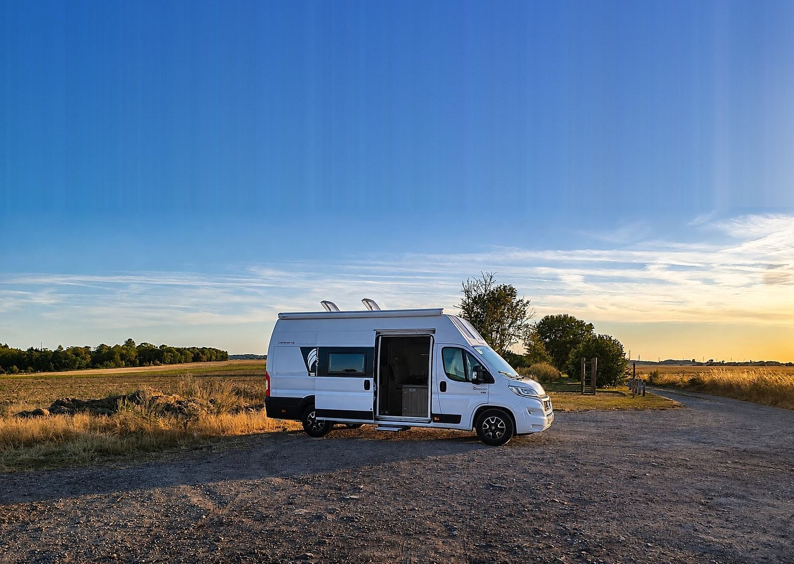 Le van aménagé Dreamer Camper Five de Pyja Van garé en bord de champ au soleil couchant, porte latérale ouverte, à louer en Brabant wallon